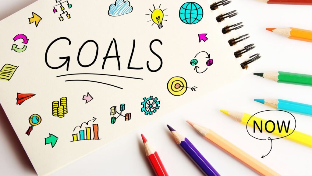 A notebook page with the word 'GOALS' surrounded by colorful hand-drawn icons, including lightbulbs, globes, arrows, and graphs, accompanied by vibrant pencils and the word 'NOW'.